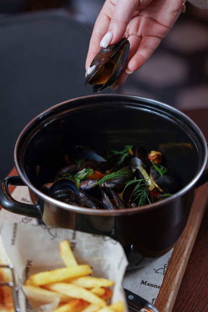 Hand holding an open cooked mussel over a pot of mussels.