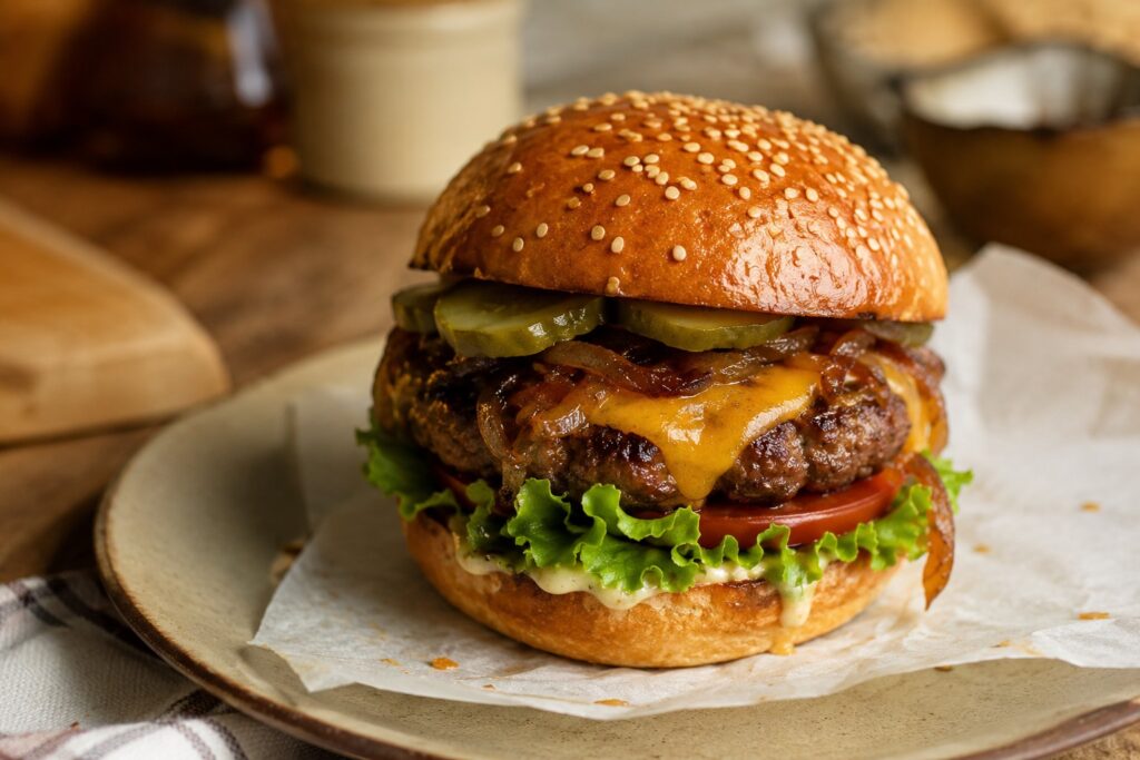 Hamburger with cheese, pickles, tomato, lettuce and mayo in a sesame seed topped bun, on a paper lined serving plate.