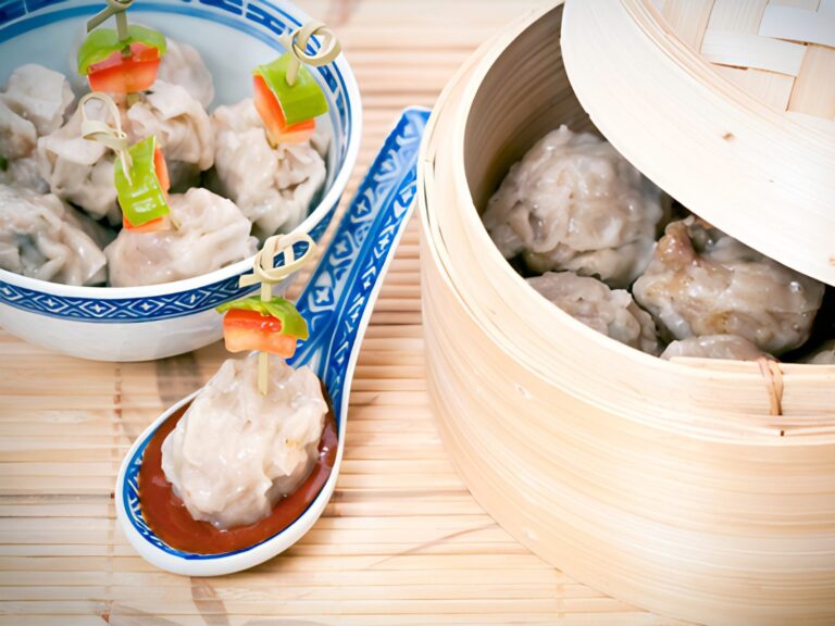 A bamboo steamer with steamed wontons, a bowl of wontons and a Chinese spoon holding a steamed wonton.