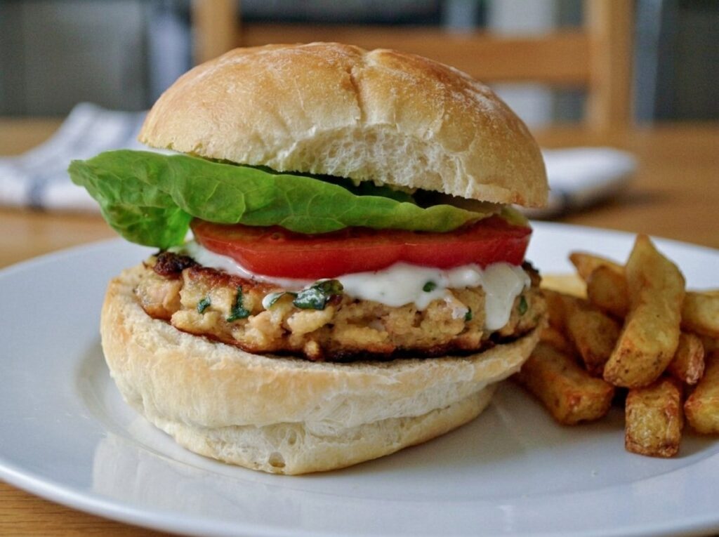 Salmon patty served in a bun with lettuce, tomato and tartar sauce, and a few fries on the plate.