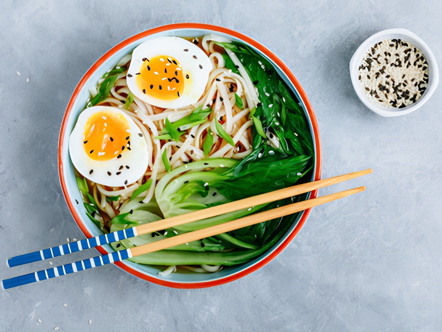 A bowl of bok choy soup with chopsticks and a small dish of sesame seeds.