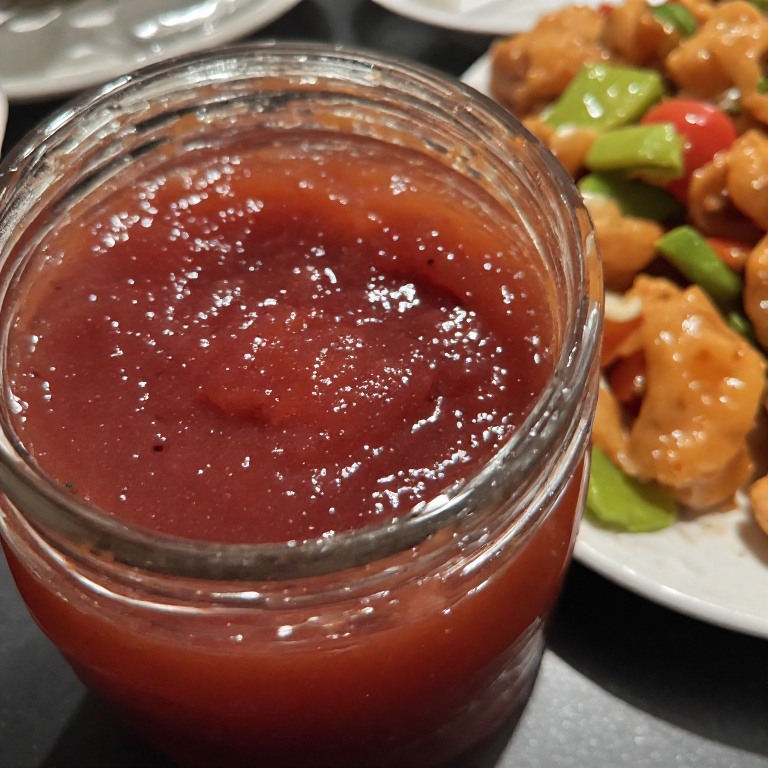 A jar of homemade plum sauce using fresh plums and some food in the background.