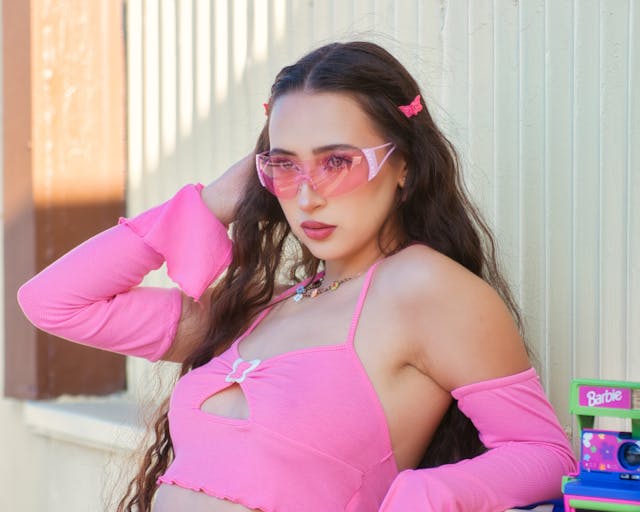 Woman in a pink outfit with pink sunglasses, standing against a wall with Barbie-themed accessories