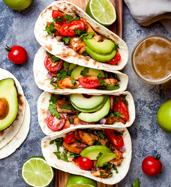 Several chicken tacos on a wooden board, with avocado and cherry tomatoes