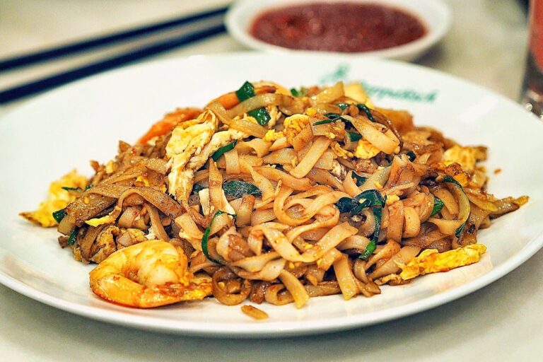 Char kway teow on a white plate with sauce and chopsticks in the background.