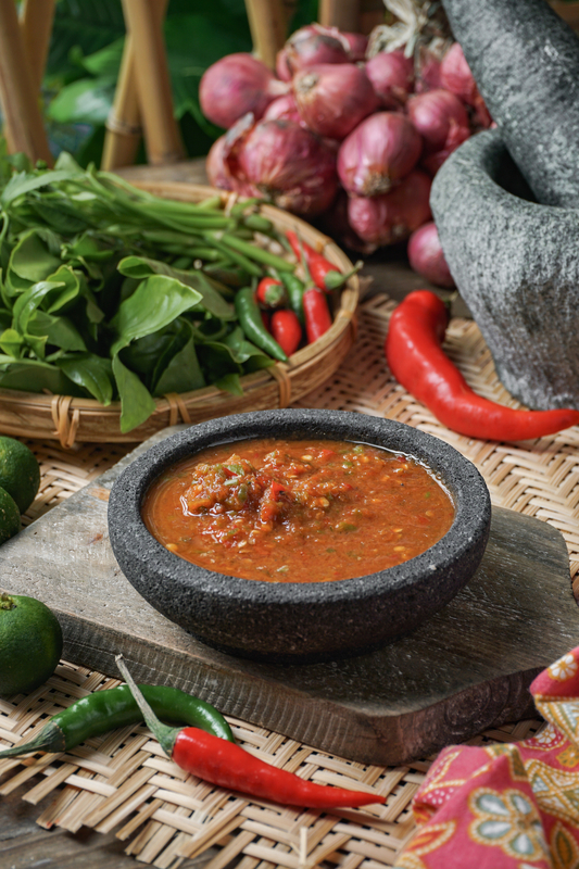 A stone bowl of homemade sambal belacan with chilies and onions in the background.