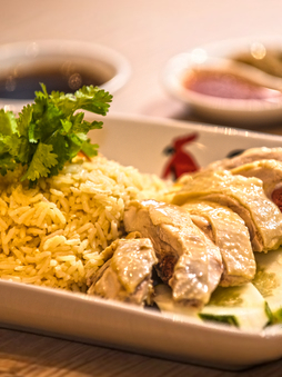 A plate of Hainanese chicken rice with fresh herb garnish, cucumber slices and 2 sauces.