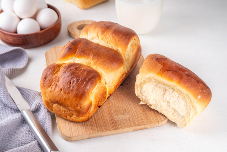 Japanese milk bread buns on a wooden board with one bun cut open to show the soft crumb.