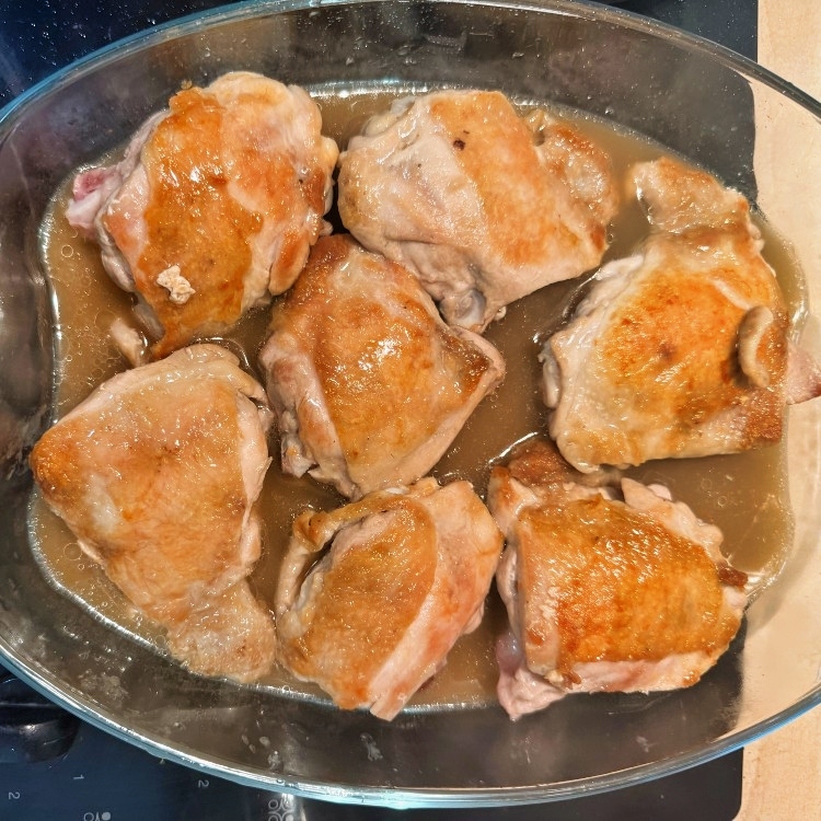 Browned chicken thighs arranged skin-side up in a glass baking dish with broth.