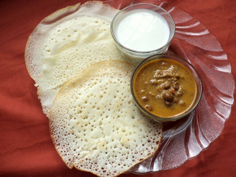 Appam with lacy edges served with kadala curry and coconut milk on a glass plate.