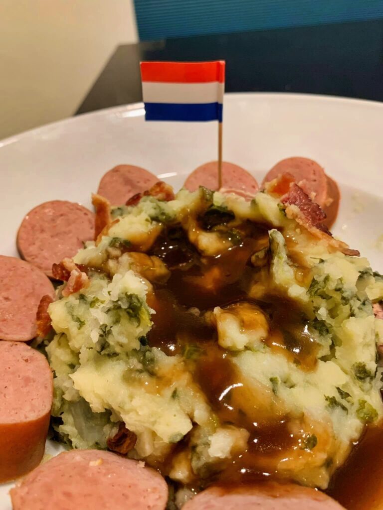 Close-up of kale stamppot with mushroom gravy, bacon, sliced smoked sausage and a Dutch flag.