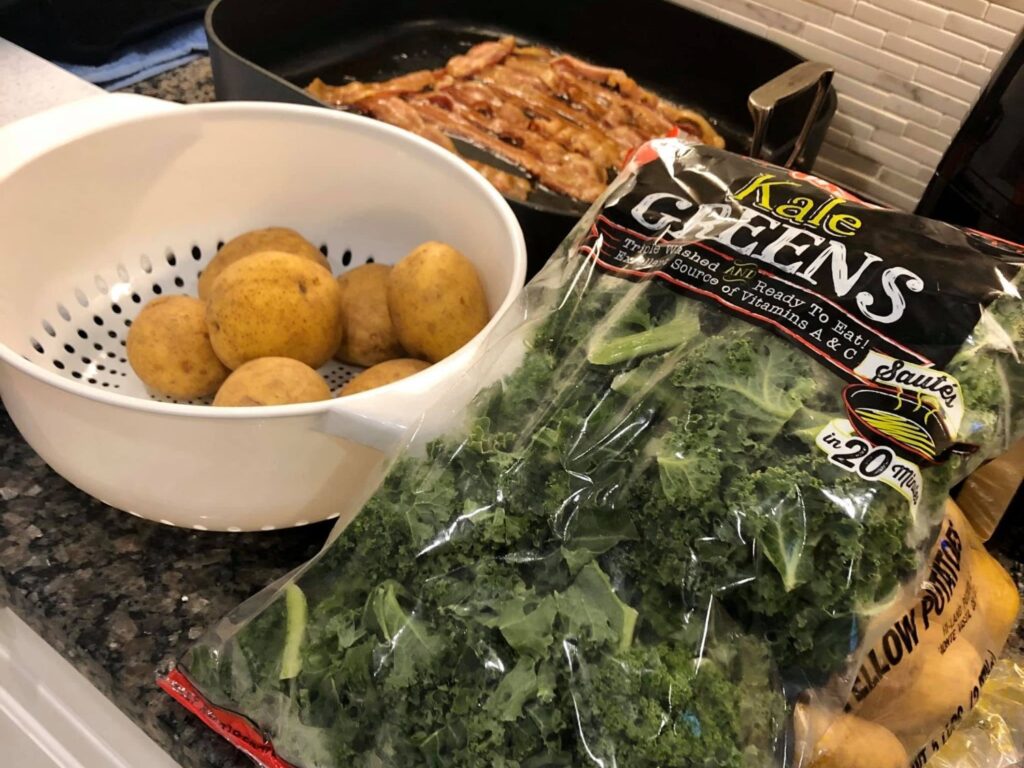 Potatoes in a colander beside a bag of chopped kale and bacon cooking in a skillet.