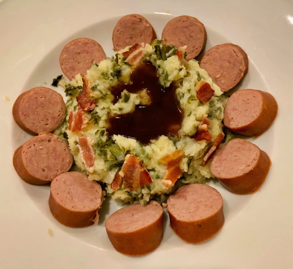 Overhead view of kale stamppot topped with bacon and brown gravy, surrounded by smoked sausage slices.