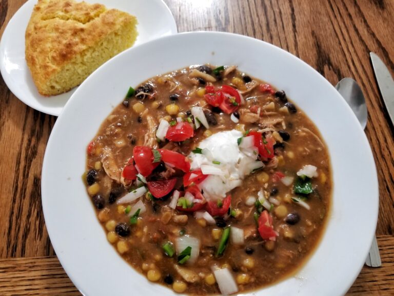 A bowl of chicken soup with beans, corn and fresh toppings, served with cornbread.
